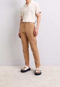 Ljusbruna bomullsh shortsbyxor, avslappnad passform, som är i kombination med en kortärmad skjorta i off-white och vita sandaler på en mörk sula. Ljust beige mattbakgrund.