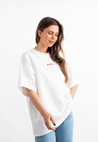 T-shirt bianca oversize in cotone, con maniche corte e stampa rossa. Abbinata a jeans in denim azzurro chiaro.