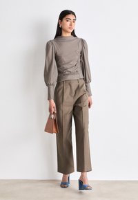 Haut long gris à manches longues avec des épaules bouffantes, associé à un pantalon marron taille haute. Des sandales à talons bleues et un petit sac à main marron complètent le look.