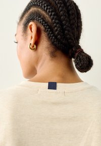 Beige t-paita pyöreällä pääntiellä, jossa on laivastonsininen etiketti takana. Mallilla on lettipäinen kampaus ja kultainen renkorengaskorvakoru.