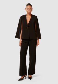 Schwarzer, taillierter Blazer mit tiefem V-Ausschnitt, Umhangärmeln und einem einzelnen Knopfverschluss, kombiniert mit schwarzen hochtaillierten Hosen und hohen Schuhen.