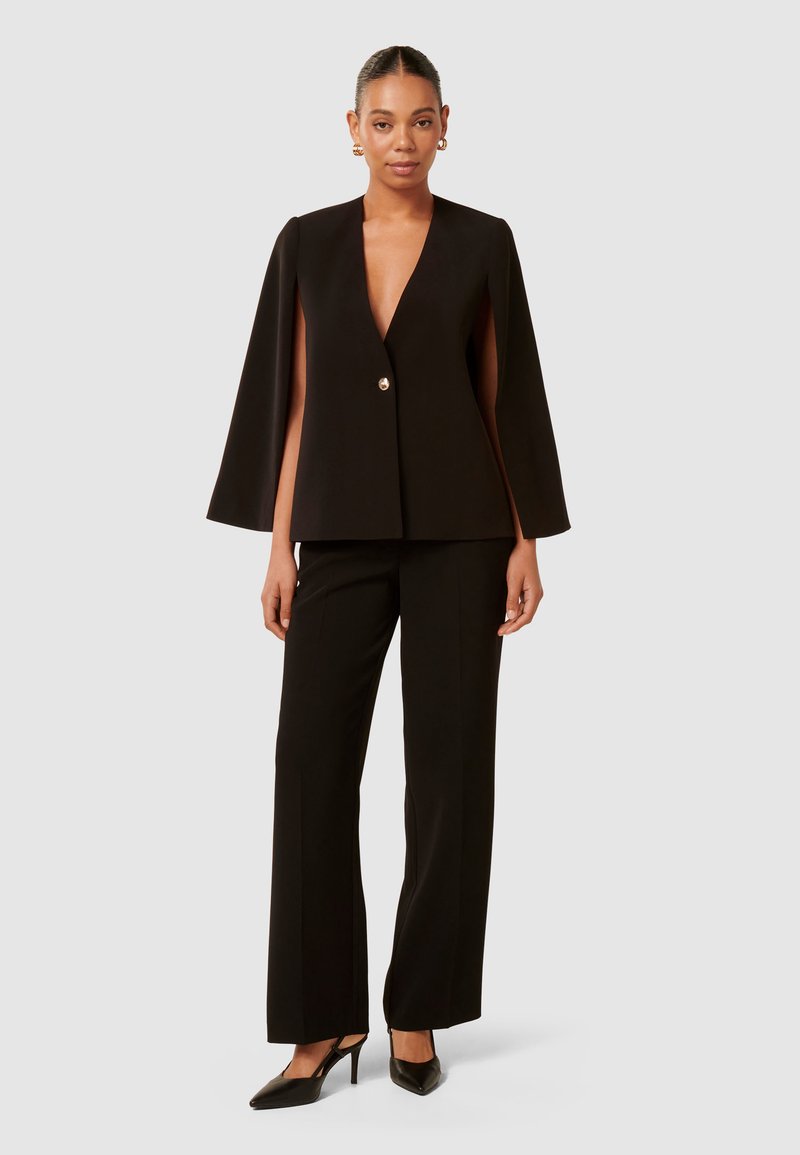 Schwarzer, taillierter Blazer mit tiefem V-Ausschnitt, Umhangärmeln und einem einzelnen Knopfverschluss, kombiniert mit schwarzen hochtaillierten Hosen und hohen Schuhen.