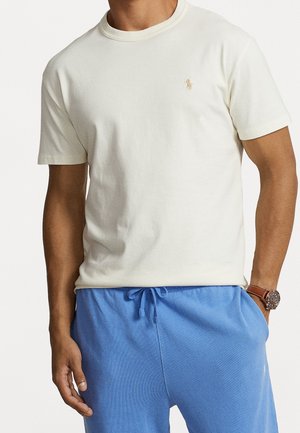 Baumwoll-T-Shirt mit Rundhalsausschnitt in Off-White, mit kurzen Ärmeln und einem subtilen Logo in Hellbraun. Kombiniert mit blauen Sweatpants und einer Uhr.