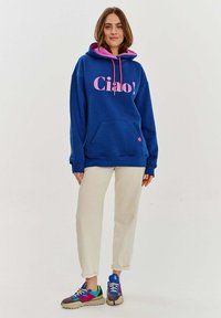 Modrá mikina s kapucí a růžovým interiérem, s nápisem "Ciao!" vytištěným velkými růžovými písmeny. Doplňují ji jednobarevné krémové kalhoty a vícebarevné tenisky.