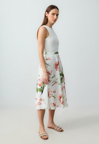 Weißes ärmelloses Oberteil kombiniert mit einem fließenden, blumigen Rock mit rosa und grünen Mustern. Das Model trägt tanfarbene Sandalen mit zwei Riemen.