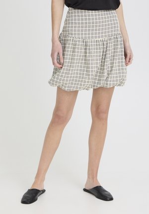 Mujer con falda burbuja de cuadros beige y negro, top negro y zapatos negros sin cordones, de pie contra un fondo claro y liso.