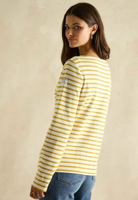 Gelb- und weiß gestreiftes Langarmshirt mit Rundhalsausschnitt, strukturierter Stoff und einem Etikett am linken Ärmel, kombiniert mit Jeans.