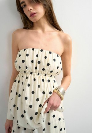 Kvinde iført hvid stropløs kjole med sorte prikker, holder stof og bærer chunky sølv- og guldbøjler på højre håndled.