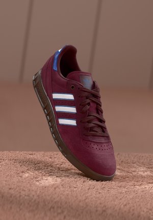 Bordowy sneaker Adidas z białymi paskami, gumową podeszwą i niebieskim detalem na pięcie unoszący się nad teksturowaną brązową powierzchnią.