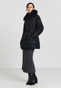 Giacca a vento nera con design trapuntato, colletto in pelliccia sintetica, chiusura con zip e vita con cintura, abbinata a gonna in maglia grigia e stivali neri con tacco.