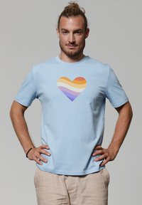 Lichtblauwe katoenen t-shirt met een hartafbeelding in oranje, geel, wit en paarse horizontale strepen. Korte mouwen en een ronde hals.