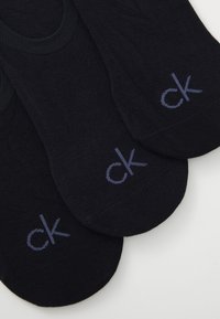 Calcetines de tobillo negros hechos de una tela suave, con una superficie texturizada y un sutil logo bordado "ck" en azul marino.