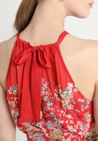 Haut rouge à bretelles nouées avec encolure froncée et détail de nœud au dos. Tissu léger présentant divers motifs floraux.