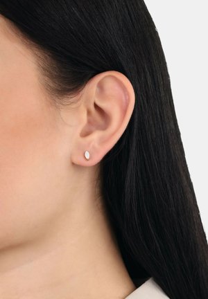 Pendientes de oro en forma de lágrima con una superficie suave y blanca, similar al ópalo, enmarcados por cabello oscuro y un fondo neutro.