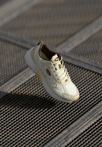 Zapatilla color crema con parte superior de cuero suave, acentos texturizados y un emblema amarillo. Suela de goma con tracción, cordones visibles en la parte frontal.
