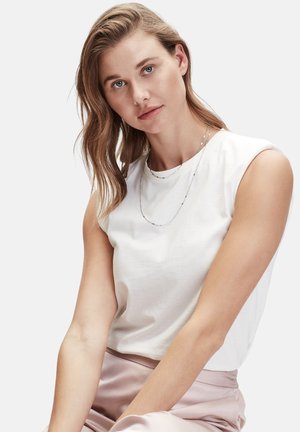 Haut sans manches en coton blanc à encolure ronde, associé à une jupe en satin rose pâle. Le modèle porte des colliers délicats en argent superposés.