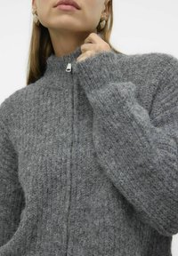 Grå strikket zip-genser med høy krage og ribbestruktur. Har lange ermer og detalj med sølvglidelås ved halsen.