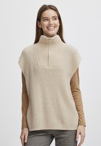 Beige bordázott kötött mellény magas gallérral, félig zipzárral és széles vállakkal, egy barna hosszú ujjú felső felett.