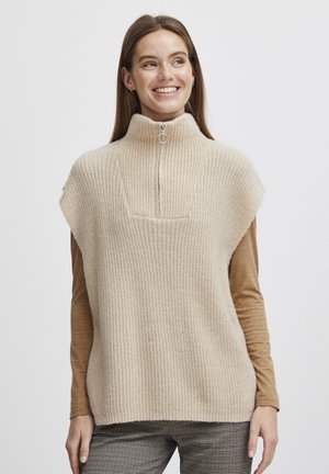 Gilet en tricot côtelé beige avec un col montant haut, présentant une fermeture éclair demi-longueur et des épaules larges, porté sur un haut à manches longues marron.