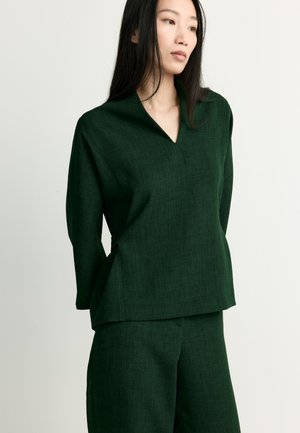 Woman with long black hair wearing a dark green textured long-sleeve top and matching pants, standing against a plain light background.