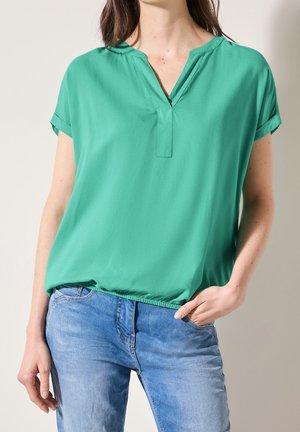 Woman wearing green short-sleeve blouse with V-neck and blue jeans, with one hand in pocket and loose hair falling over shoulders.