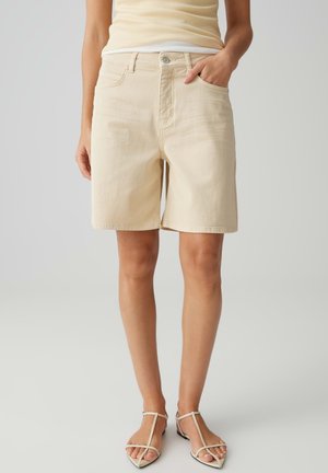 Frau trägt beige knielange Shorts, ein passendes Tanktop und beige flache Sandalen mit Riemen, steht mit einer Hand in der Tasche.
