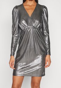 Metallisk silverfärgad klänning med långa puffärmar, en djup V-ringning och en insvept design. Har en strukturerad, glansig tyg och rynkdetalj vid midjan.