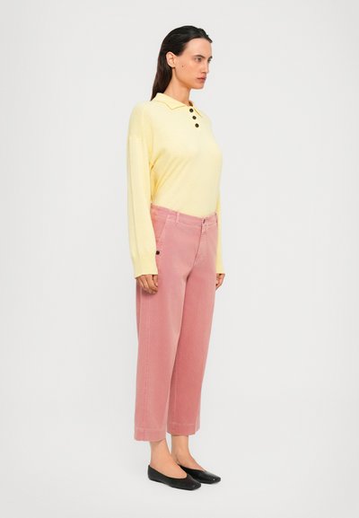 Woman standing sideways wearing a pale yellow long-sleeve polo shirt, pink wide-leg cropped pants, and black flat shoes.