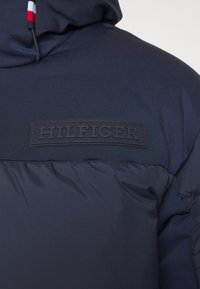 Marineblå puffjakke med en teksturert finish, med en fremtredende svart preget "HILFIGER" logo og en hette med snøring.