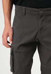 Grijze cargobroek van duurzaam materiaal, met een knoopsluiting, zijzakken en versterkte stiksels. Close-up van de taille en zak.
