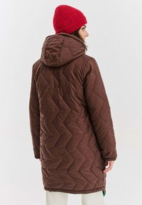 Hnedý prešívaný dlhý kabát s kapucňou, ktorý má textúrovaný vzor v tvaru cik-cak. Nosí sa s červenou pletenou čiapkou.