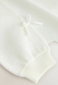 Weißer Pullover mit strukturierter Oberfläche, versehen mit einer Schleifenverzierung am Ärmel und einem gerippten Bündchen. Das Design ist weich und minimalistisch.