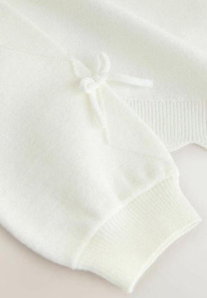 Biały sweter o fakturowanym wykończeniu, z detalem kokardki na rękawie i ściągaczem przy mankiecie. Projekt jest miękki i minimalistyczny.