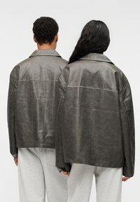 Υπερμεγέθη δερμάτινο μπουφάν ανθρακί γκρι με ίσιο γιακά και ορατές ραφές, φορεμένο πάνω από ανοιχτό γκρι φόρμες. Λείο υφή και χαλαρή εφαρμογή.