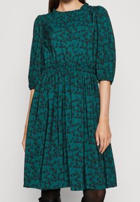 Tummansininen mekko, jossa on kerrattu vyötärö, kyynärpituinen hihna ja röyhelökaula-aukko. Mekossa on musta ja tummanvihreä kukkakuosi.