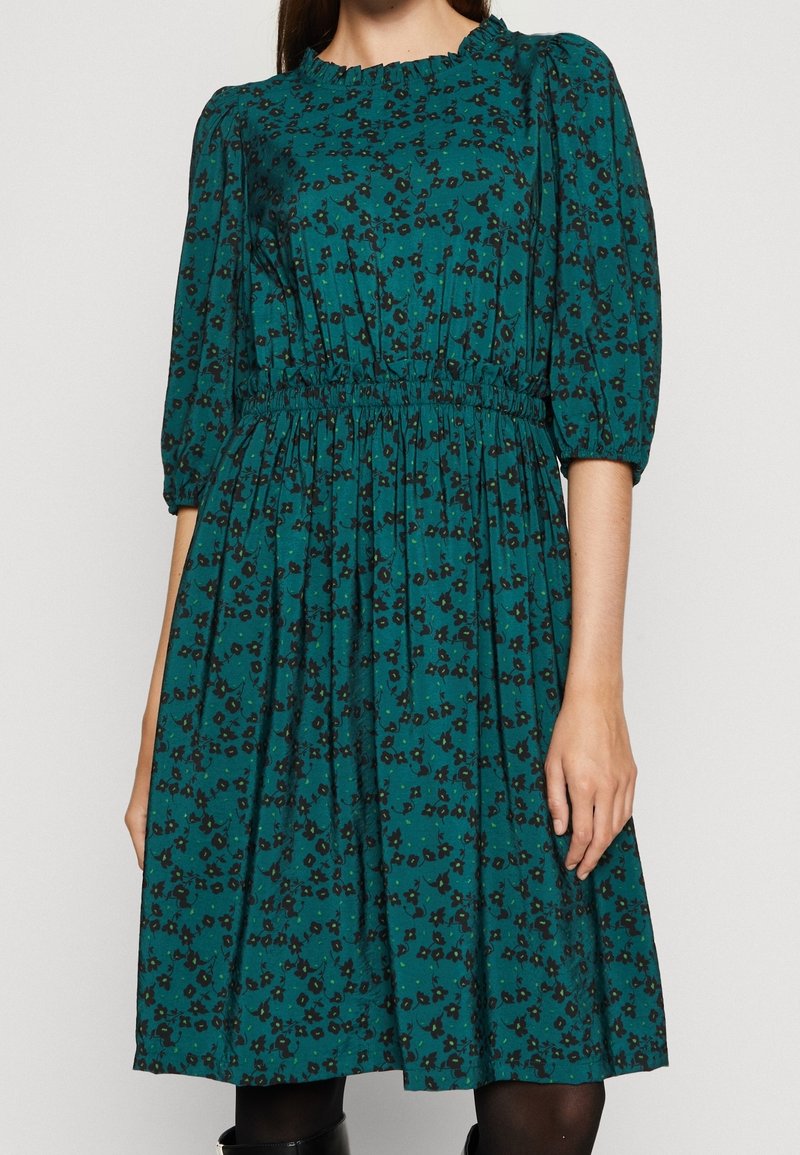 Tummansininen mekko, jossa on kerrattu vyötärö, kyynärpituinen hihna ja röyhelökaula-aukko. Mekossa on musta ja tummanvihreä kukkakuosi.