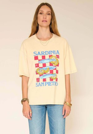 Femme portant un T-shirt beige ample avec un motif de poisson bleu et de citron et un motif à damier rouge, avec le texte "Sardaigne San Pietro", assorti à un jean bleu.
