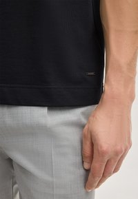 Zwart katoenen T-shirt met een afgeronde zoom en een klein metalen merketiket. Gecombineerd met lichtgrijze, gestructureerde broek.