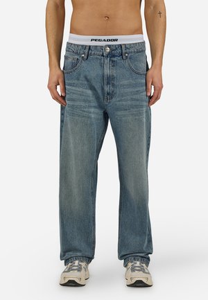 Mann iført vide blå denimjeans, hvit undertøy med synlig merkelogo på midjebåndet og beige joggesko, stående mot en enkel bakgrunn.