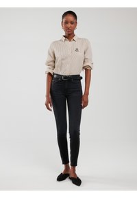 Tânără în picioare, purtând o cămașă bej cu dungi și nasturi, curea neagră, blugi skinny negri și pantofi slip-on negri, pe fundal alb.