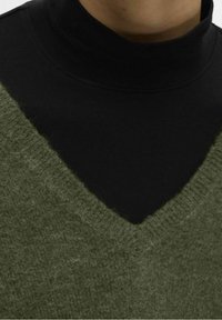 Maglione in maglia verde oliva con scollatura a V indossato sopra un dolcevita nero. Materiale morbido e testurizzato con dettagli di cucitura visibili.