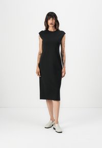 Mustahdim svartärndress uden ærmer med en lige skæring, knælangt design, lavet af glat stof, parret med lysegrå slip-on sko.