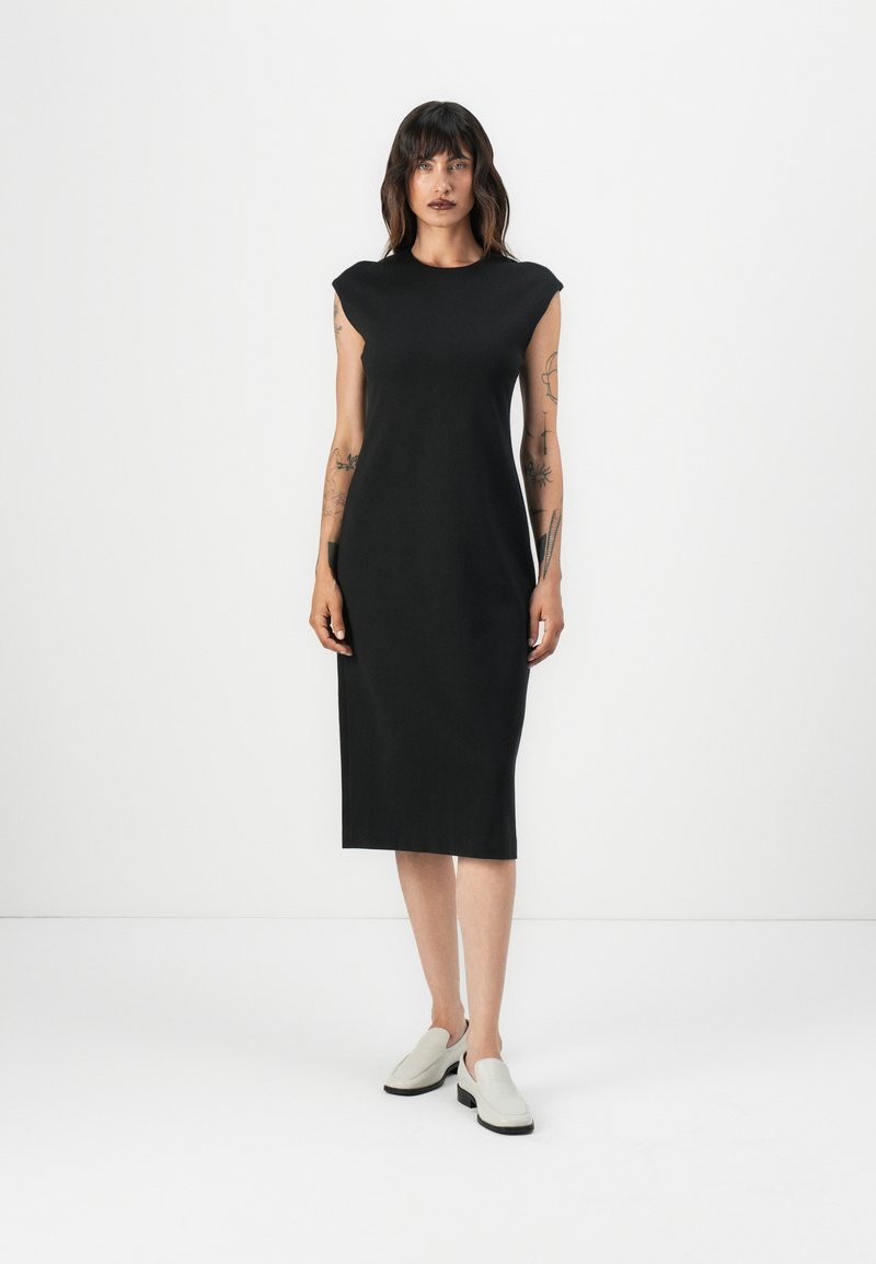 Mustahdim svartärndress uden ærmer med en lige skæring, knælangt design, lavet af glat stof, parret med lysegrå slip-on sko.