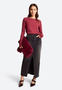 Burgunderfarbener Langarm-Top kombiniert mit einem schwarzen Denim-Maxi-Rock mit einem Schlitz vorne. Das Model hält eine burgunderfarbene Faux-Fur-Tasche und trägt spitze Absätze.