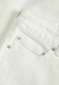 Il tessuto di denim bianco mostra un primo piano di una tasca, con accenti di bottoni argentati e una superficie testurizzata con dettagli di cucitura visibili.