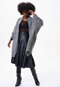 Cardigan en tricot gris surdimensionné avec des poignets côtelés, associé à un haut noir transparent et à une jupe en cuir noir taille haute, porté avec des bottes montantes.