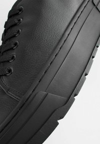 Adidași negri din piele, cu un superior texturat, talpă groasă din cauciuc și șireturi plate. Vederea laterală evidențiază detaliile cusăturii și designul minimalist.