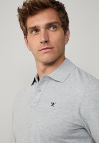 Harmaa poolopaita, jossa kaulus, tummansinisiä yksityiskohtia ja pieni logo. Materiaali vaikuttaa pehmeältä ja kevyeltä, klassinen istuvuus.