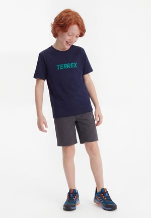 Băiat roșcat, purtând un tricou TERREX bleumarin, pantaloni scurți gri și adidași de drumeție albaștri, stând într-o postură zâmbitoare și relaxată.