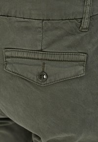 Kapsa na kalhotách z olivově zelené látky s diagonálním střihem, zapínáním na jeden knoflík a kontrastním prošitím podél okrajů.